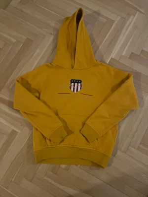Senapsgul GANT hoodie - Snygg senapsgul hoodie med broderat GANT tryck och emblem på bröstet. Hoodien har en stor huva, ribbade muddar vid ärmar och nederkant samt en avslappnad passform. Perfekt för dig som gillar streetwear och vill sticka ut med färg.