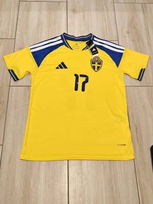 Sverige matchtröja Gyökeres #17 - Gul och blå matchtröja från Adidas för Sveriges landslag med Gyökeres och nummer 17 på ryggen. Tröjan har korta ärmar, blå detaljer på axlar och ärmslut samt broderad landslagssköld på bröstet. Tillverkad i lätt och ventilerande material.