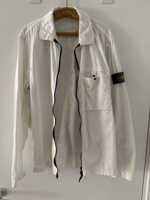 Vit overshirt från Stone Island - Snygg vit overshirt från Stone Island med dragkedja framtill, bröstficka och klassisk logopatch på ärmen. Overshirten har en avslappnad passform och är tillverkad i ett lätt material som passar perfekt till lager-på-lager-stil.
