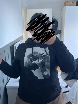 Svart hoodie med Tupac-tryck - Säljer en svart hoodie med stor vit/svart Tupac-tryck på bröstet. Hoodien har huva med dragsko och en klassisk känguruficka framtill. Materialet känns mjukt och bekvämt, perfekt för en avslappnad streetwear-look. Fickan har även en liten öppning som man ser på bilden men det är inget som man märker av