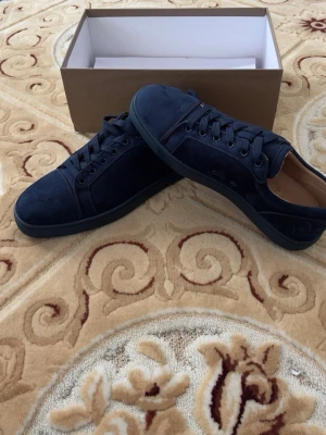 Mörkblå sneakers med röd sula - Snygga mörkblå sneakers från Christian Louboutin i mocka med klassisk röd sula och svarta skosnören. Skorna har en stilren design med lågt skaft och diskreta detaljer. Perfekta för dig som vill sticka ut med exklusiva detaljer.