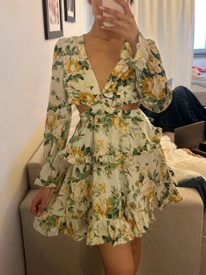 Blommig volangklänning med cut-outs - Supersöt kort klänning med volanger och blommigt mönster i gult, grönt och vitt. Djup v-ringning, långa ärmar och trendiga cut-outs i midjan. Oanvänd! 