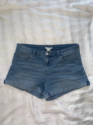 Blå denimshorts med uppvik - Säljer ett par klassiska blå denimshorts med uppvikta benslut och femficksmodell. Shortsen har normal passform och stängs med knapp och dragkedja. Perfekta för varma dagar och enkla att matcha med olika toppar.
