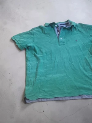 Grön piké från Tommy Hilfiger - Säljer en grön pikétröja från Tommy Hilfiger med korta ärmar och klassisk krage. Tröjan har knappar vid halsen och en liten logga på bröstet. Perfekt för en avslappnad och stilren look.