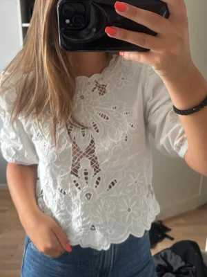 Vit blus med broderade spetsdetaljer - Säljer en vit blus med puffärm och broderade blommor samt spetsdetaljer framtill. Blusen har rundad halsringning och vågig kant nertill. Perfekt för dig som gillar romantisk och söt stil. Någon tråd har gått av i mitten på tröjan men de går enkelt att fixa 