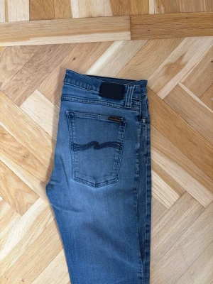 Blå Nudie Jeans - Snygga blå jeans från Nudie Jeans med klassisk femficksdesign och diskret slitning på låren. Jeansen har en mörkare tvätt och den ikoniska vågade sömmen på bakfickan. Perfekta för en avslappnad och trendig look.