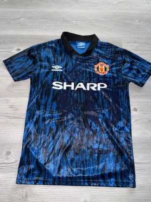 Manchester United retro bortatröja - Säljer en klassisk Manchester United bortatröja från Umbro i blått och svart mönster med svart krage. Tröjan har klubbmärke på bröstet och SHARP som sponsortryck. Perfekt för dig som älskar vintage fotbollströjor.