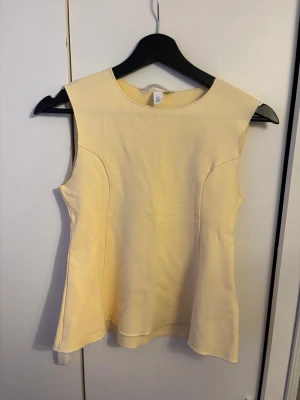 Gul ärmlös topp - Snygg ljusgul ärmlös topp med rund halsringning och figurnära passform. Toppen har en lätt utsvängd nederkant och är sydd i ett mjukt, slätt tyg. 