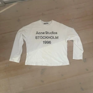 Vit långärmad Acne Studios t-shirt - Vit långärmad t-shirt från Acne Studios med tryck i svart text på bröstet där det står 'Acne Studios STOCKHOLM 1996'. Klassisk passform och rund halsringning. Perfekt för en clean och stilren look. Finns en liten fläck på insida arm som inte syns vid användning.