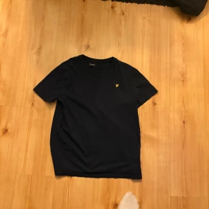 Marinblå t-shirt från Lyle & Scott - Snygg marinblå t-shirt från Lyle & Scott med klassisk rund hals och gul broderad logga på bröstet. Tillverkad i mjuk bomull och har en normal passform. Perfekt basplagg för alla tillfällen.