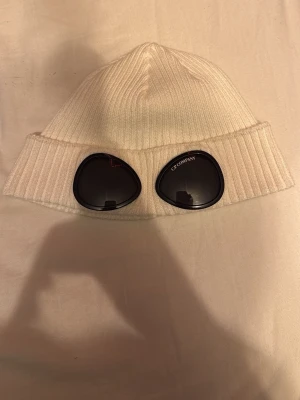 Vit ribbstickad mössa från cp company  - Snygg vit ribbstickad mössa med uppvikt kant. Klassisk och enkel design som passar till de flesta outfits. Perfekt för kalla dagar när du vill hålla dig varm och stilren. Äkta cp company mössa med glasögon med en liten skråma på baksidan 
