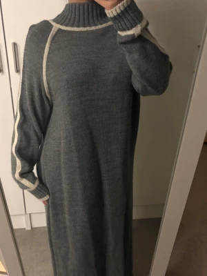 Grå stickad långklänning med vita detaljer - Mysig grå stickad långklänning med vita kontrastsömmar längs ärmar och axlar. Klänningen har hög krage och långa ärmar med ribbade muddar. Perfekt för kalla dagar och enkel att styla med sneakers eller boots.