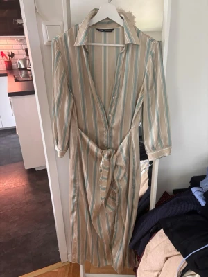 Randig långklänning med knytband - Snygg långklänning med vertikala ränder i beige, vitt och ljusblått. Klänningen har krage, knappar framtill, trekvartsärm och ett matchande knytband i midjan. Perfekt för en avslappnad men stilren look.