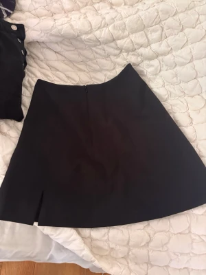 Svart A-linjeformad kjol - En svart, stilren kjol i A-linjeform med diskret slits framtill. Kjolen har en enkel design utan synliga detaljer och är gjord i ett slätt tyg som ger en klassisk look. Perfekt att matcha med olika toppar för en trendig outfit.