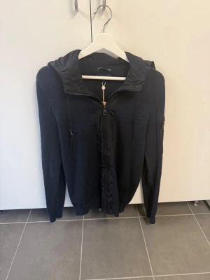 Mörkblå cardigan från Salsa, Stl M - Mörkblå cardigan från Salsa med dragkedja framtill och justerbar huva. Tillverkad i stickat material med detaljer i tyg längs dragkedjan och huvan. Långa ärmar och ficka på ena ärmen ger en cool touch. Perfekt för en avslappnad stil.
