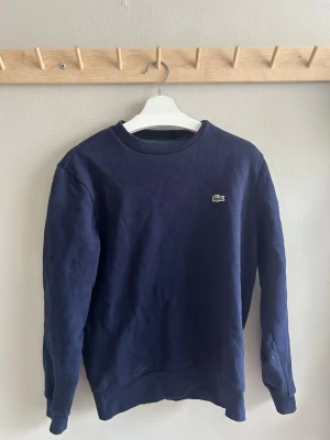 Mörkblå sweatshirt från Lacoste - Klassisk mörkblå sweatshirt från Lacoste med rund halsringning och den ikoniska krokodilloggan broderad på bröstet. Tröjan har ribbade muddar vid ärmslut och nederkant. Perfekt för en avslappnad och stilren look. Köptes på best secret !!