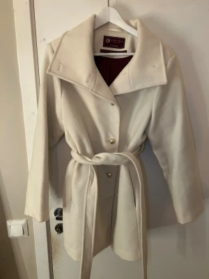 Krämvit kappa från Amisu - Stilren krämvit kappa från Amisu med bred krage, knytskärp i midjan och knappar framtill. Kappan har ett mjukt yttre och vinrött foder. Perfekt för kyliga dagar och ger en elegant look till din outfit.