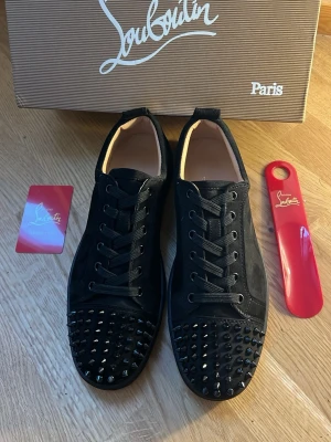 Svarta sneakers med nitar från Louboutin - Snygga svarta sneakers från Christian Louboutin i mocka med svarta nitar på tån och sidorna. Skorna har klassiska svarta skosnören och den ikoniska röda sulan. Perfekta för dig som vill sticka ut med en edgy och lyxig stil.