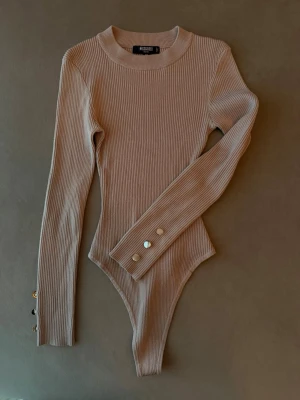 Beige ribbad body med guldknappar - Beige/brun ribbstickad body från Missguided med lång ärm och rund hals. Snygga guldfärgade knappar på ärmsluten, de på högra sidan har fått lite missfärgning. 