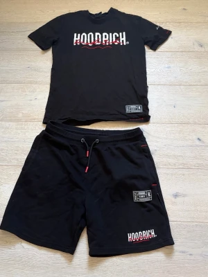 Svart Hoodrich set med t-shirt & shorts - Säljer ett svart set från Hoodrich med matchande t-shirt och shorts. Båda plaggen har vita och röda logotexter samt detaljer som dragsko i midjan på shortsen. Materialet ser ut att vara mjukt bomullstyg, perfekt för en avslappnad stil.
