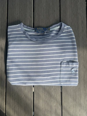Blåvit randig t-shirt Polo Ralph Lauren - Snygg blå t-shirt med vita horisontella ränder från Polo Ralph Lauren. Klassisk passform med korta ärmar, rund halsringning och en liten bröstficka med broderad logga. Perfekt för en avslappnad och stilren look.