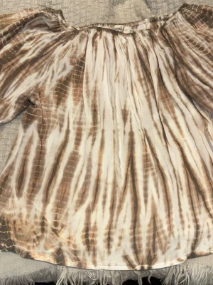 Brun batikblus från Coconut Milk - Säljer en luftig batikmönstrad blus från Coconut Milk i bruna och beige toner. Blusen har offshoulder-modell och vida, långa ärmar. Materialet känns tunt och flowy, perfekt för en avslappnad stil. Elastisk kant upptill för skön passform.