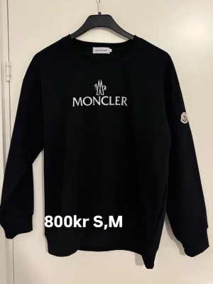 Svart Moncler hoodie med logga - Svart hoodie från Moncler med vit broderad logga på bröstet och Moncler-märke på ärmen. Klassisk passform med rund hals och ribbade muddar. Perfekt för dig som gillar stilrena och exklusiva plagg.
