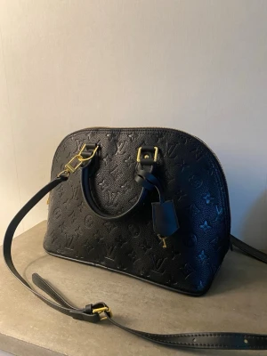 Svart handväska i läder - Snygg svart handväska  i präglat läder med klassiskt monogrammönster. Väskan har guldfärgade metalldetaljer, dubbla handtag och avtagbar axelrem. Perfekt för dig som vill ha en lyxig och tidlös accessoar.