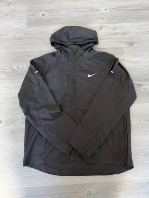 Svart vindjacka från Nike med huva - Snygg svart vindjacka från Nike med dragkedja framtill och huva. Jackan har en vit Nike-logga på bröstet och är tillverkad i ett lätt, vindtätt material. Perfekt för blåsiga dagar och enkel att matcha med olika outfits.