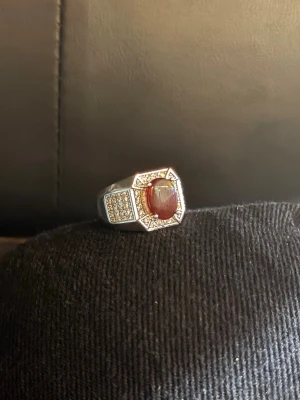 Silverring med röd sten och kristaller - Snygg silverfärgad ring med en stor röd oval sten i mitten, omgiven av små gnistrande kristaller . Ringen har en bred design och fyrkantig front med extra kristalldetalijer på sidorna. Perfekt för dig som gillar statement-smycken och vill sticka ut. 925 silver stämpel 