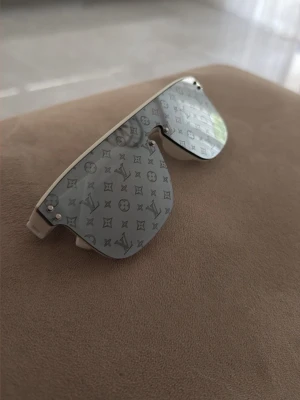 Silverfärgade monogram solglasögon - Säljer ett par riktigt snygga solglasögon med spegelglas i silver och monogrammönster över hela linsen. Bågarna är vita och modellen har en futuristisk, sköldformad design som sticker ut. Perfekta för dig som vill ha en unik accessoar till sommaren.