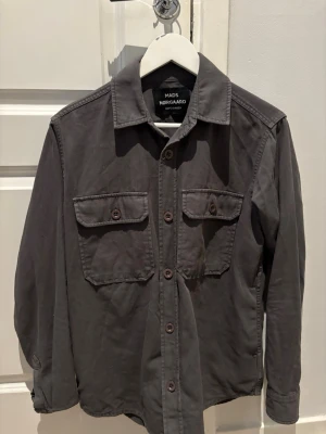 Mörkgrå overshirt från Mads Nørgaard - Stilren mörkgrå overshirt från Mads Nørgaard med två stora bröstfickor med knappar och klassisk krage. Skjortan har knappstängning framtill och är tillverkad i ett mjukt bomullstyg. Perfekt lager-på-lager-plagg för en avslappnad look.