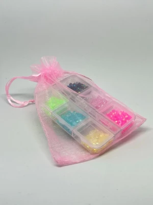 Mini kit pärlor i ask - Säljer ett set med små, färgglada plastpärlor i olika nyanser som rosa, blå, gul, grön och svart. Pärlorna kommer i en genomskinlig plastask med flera fack och levereras i en söt rosa organzapåse. Perfekt för att skapa egna smycken eller accessoarer.