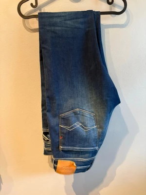 Mörkblå slim fit jeans Replay - Säljer ett par mörkblå slim fit jeans med snygg tvätt och kontrastsömmar. Jeansen har klassiska fem fickor och en läderpatch bak i midjan. Perfekta för dig som gillar en modern och stilren look.