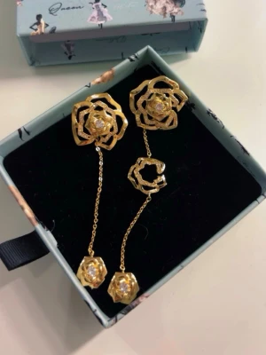 IOAKU örhängen-guld - Unika örhängen i guldfärgad metall med rosformade detaljer och hängande kedjor. Örhängena har en modern, asymmetrisk design och gnistrande stenar i mitten av varje ros. Perfekta för dig som vill sticka ut med en trendig accessoar. Nypris 599kr, aldrig använt.