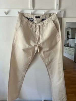 Beige chinos med avslappnad passform - Snygga beige chinos med rak passform och klassiska sidfickor fram samt två bakfickor med knapp. Tillverkade i ett mjukt bomullstyg som känns skönt mot huden. Perfekta för en clean och stilren look.