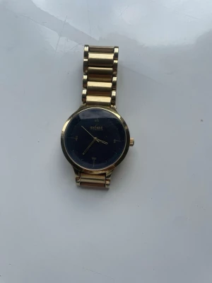 Guldfärgad klocka från Axcent - Stilren guldfärgad klocka från Axcent med metallarmband och svart urtavla. Klockan har en klassisk rund form och detaljer i guld. Perfekt accessoar för dig som vill ha en elegant och tidlös look😊.