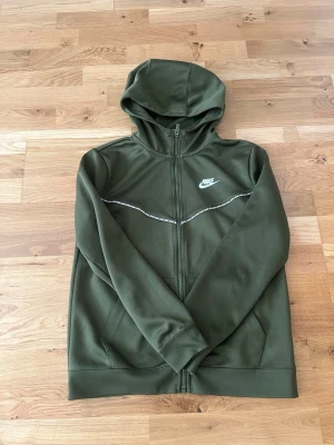 Olivgrön Nike hoodie med dragkedja - Snygg olivgrön hoodie från Nike med hel dragkedja framtill och vit logga på bröstet. Hoodien har huva, två fickor och en tunn vit detaljlinje över bröstet. Tillverkad i mjukt och bekvämt material, perfekt för en avslappnad stil.