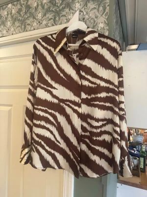 Brun och vit zebramönstrad skjorta - Elegant skjorta i glansig satinkvalitet med ett mjukt ikat-inspirerat zebra­mönster i bruna och vita toner. Den har en avslappnad oversizepassform med klassisk krage, knäppning framtill, långa ärmar med manschetter och en rundad nederkant. Ett stilrent plagg som ger en lyxig känsla både till vardags och mer uppklädda tillfällen.