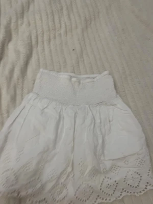 Vit kortkjol med broderad spets från Gina Tricot - Söt vit kortkjol från Gina Tricot med bred smock i midjan och fina broderade spetsdetaljer längst ner. Kjolen har en luftig och somrig känsla, perfekt för varma dagar. Passar dig som gillar en feminin och avslappnad stil.