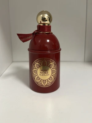 Guerlain Musc Noble Eau de Parfum - Lyxig parfym från Guerlain i en elegant mörkröd flaska med gulddetaljer och dekorativt mönster. Flaskan har en rundad form, guldfärgat klotlock och en röd rosett vid halsen. Doften heter Musc Noble och är en Eau de Parfum.