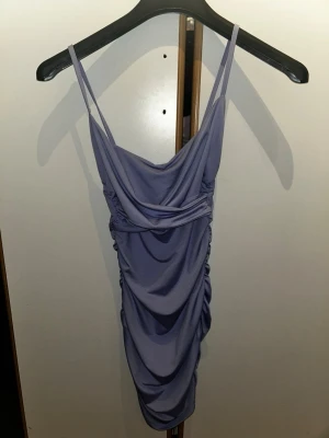 Ljuslila omlott kortklänning - Säljer en snygg ljuslila kortklänning med tunna axelband och draperad omlottdesign. Klänningen har rynkade detaljer längs sidorna och en mjuk, följsam passform. Perfekt för dig som vill sticka ut med en trendig look.
