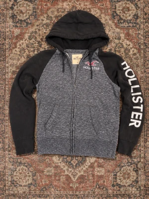 Grå och svart Hollister hoodie - Snygg zip-hoodie från Hollister i grått med svarta ärmar och huva. Stor logga på ena ärmen och mindre logga med fågel på bröstet. Mjuk bomullsblandning, två fickor fram och dragsko i huvan. Perfekt för en avslappnad och trendig look.