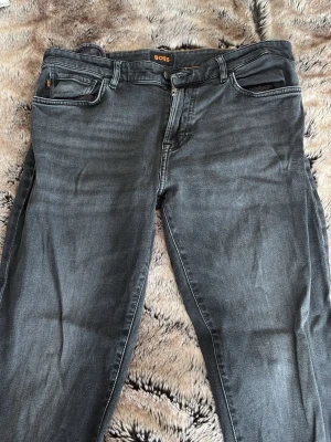 Mörka jeans från BOSS - Snygga gråa jeans från BOSS med klassisk femficksmodell och raka ben. Jeansen har subtila slitningar och en diskret logga på bakfickan. Tillverkade i mjukt denimtyg som ger en skön passform och stilren look.