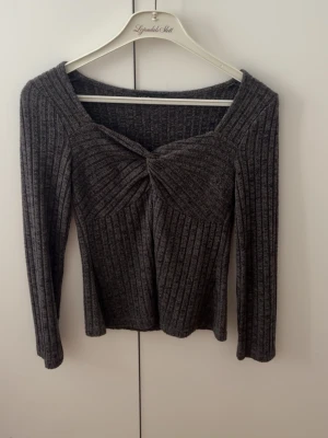 Mörkgrå ribbad topp med twist - Snygg mörkgrå ribbad topp med lång ärm och twistad detalj framtill. Toppen har en smickrande v-ringning och är tillverkad i ett mjukt, stretchigt material som sitter bekvämt på kroppen. Perfekt för dig som gillar stilrena plagg med det lilla extra. LITEN I STORLEKEN