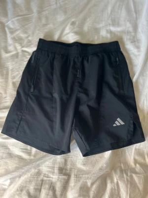 Svarta träningsshorts från Adidas - Snygga svarta träningsshorts från Adidas med elastisk midja och två dragkedjeförsedda fickor. Shortsen har en diskret Adidas-logga på ena benet och är tillverkade i ett lätt och snabbtorkande material, perfekta för aktiva dagar. Rätt så stor i storleken
