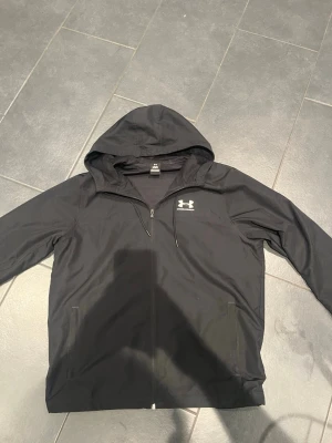 Svart hoodie från Under Armour - Snygg svart hoodie från Under Armour med dragkedja framtill och justerbar huva. Tillverkad i ett lätt och sportigt material med meshfoder på insidan. Logga i vitt på bröstet och två fickor på sidorna. Perfekt för en aktiv livsstil.