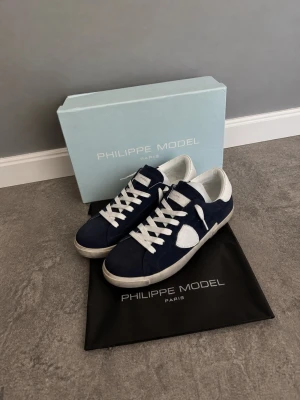 Philippe Model PRSX LOW Mörkblå suede  - Snygga sneakers från Philippe Model i marinblå mocka med vita detaljer och vita skosnören. Skorna har en klassisk låg siluett och vit sula. Perfekta för dig som gillar stilrena och trendiga sneakers med exklusiv känsla. Dustbag medföljer.