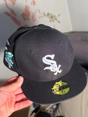 Svart White Sox keps 59FIFTY - Snygg svart keps från New Era 59FIFTY med White Sox-logga broderad framtill. På sidan finns broderi med texten 'Greetings from Chicago' och en grön vägskylt med 'Chicago Southside'. Klassisk rak skärm och strukturerad passform.