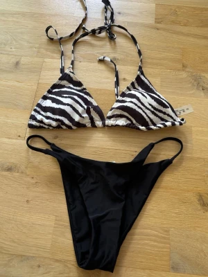 Zebramönstrad bikini med svart underdel - Snygg bikini med trekantsöverdel i zebramönster och knytband i nacken och ryggen. Underdelen är enkel och svart med smala band i sidorna. Underdelen är aldrig använd, toppen är använd men endast några gånger, liten brun fläck på den som man kan se på sista bilden men det går säkert bort i tvätten